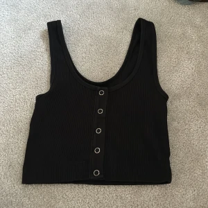 Svart ribbad croppad linnetopp från BikBok - Säljer en svart ribbad croppad linnetopp från BikBok med dekorativa silverfärgade knappar framtill. Toppen har breda axelband och är perfekt för sommaren. Enkel och stilren design som passar till mycket.