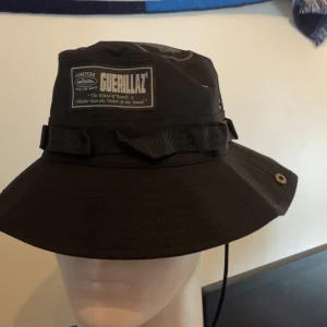 Svart bucket hat från Cortiez - Svart bucket hat från Cortiez med patch framtill och tryckt motiv på ovansidan. Hatten har dekorativa band runtom och metallöljetter på sidorna. Materialet är en mix av bomull och polyester.