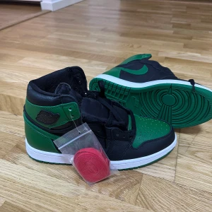 Nike Air Jordan 1 Retro High svart och grön - Säljer ett par Nike Air Jordan 1 Retro High i svart och grönt med vit sula. Skorna har klassisk snörning och rund tå. Extra röda skosnören medföljer. Perfekta för dig som gillar streetwear och sneakers med ikonisk design. Storlek 43