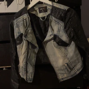 Svart och ljusblå bikerinspirerad jeansjacka - Säljer en cool jeansjacka från Pull&Bear i ljusblå denim med svarta skinnliknande detaljer på axlar och ärmar. Jackan har dragkedja, knappar och två fickor framtill. Perfekt för dig som vill sticka ut med en edgy stil.