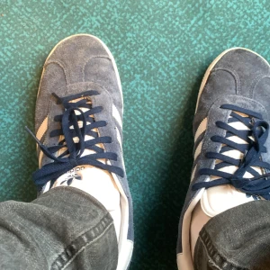 Adidas Gazelle blå sneakers - Säljer ett par klassiska Adidas Gazelle sneakers i blå mocka med vita detaljer och snörning. Skorna har den ikoniska tre-randiga designen på sidorna och platt sula. Perfekta för dig som gillar retrostil!