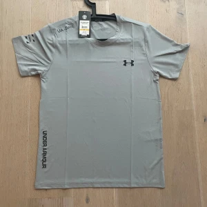 Grå t-shirt från Under Armour - Snygg grå t-shirt från Under Armour med svart logga på bröstet och text längs sidan.  Helt nya med tag och aldrig använd .