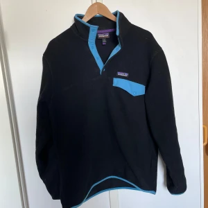 Svart fleece-tröja från Patagonia - Svart fleece-tröja från Patagonia med blå detaljer vid krage, knappslå och bröstficka. Tröjan har lång ärm och en klassisk passform, perfekt för kyligare dagar. Ikonisk Patagonia-logga på bröstet.