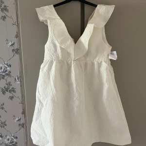 Vit Vera klänning med volangdetalj - Supersöt vit klänning från bubbleroom med djup v-ringning och breda axelband med volangdetaljer. Klänningen har en lätt strukturerad yta och är ärmlös, perfekt sommaren eller studenten, säljer den då jag tyvärr fick hem fel storlek, helt ny med prislapp kvar🌸🩷 