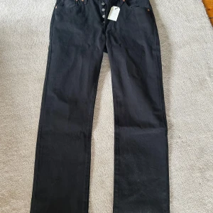 Svarta Levi's 501 jeans W32 L34 - Klassiska svarta Levi's 501 jeans med raka ben och knäppning fram. Tillverkade i 100% bomull och har den ikoniska läderpatchen bak i midjan. Passar dig som gillar tidlös stil och vill ha ett par jeans som funkar till allt. Helt oanvända, säljer pga dom var för små.