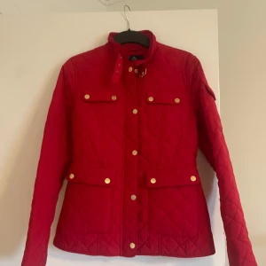 Röd quiltad jacka från Stockh LM - Säljer en snygg röd quiltad jacka från Stockh LM med guldiga knappar och hög krage. Jackan har fyra fickor framtill och markerad midja för en stilren look. Perfekt för dig som vill sticka ut med färg i vardagen.