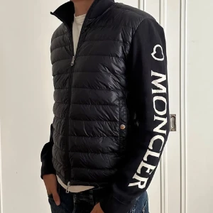 Moncler cardigan - Mörkblå cardigan från Moncler med stora vita logotyptryck på ena ärmen. Jackan har quiltad framsida i glansigt material, dragkedja och två fickor med knappar. Ärmarna är i mjukare tyg och jackan har hög krage.