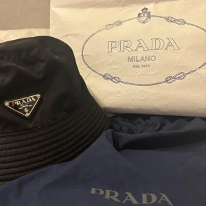 Svart bucket hat från Prada - Säljer en svart bucket hat från Prada i återvunnen nylon med klassisk logga framtill. Hatten har en stilren design och är perfekt för att lyfta vilken outfit som helst. 