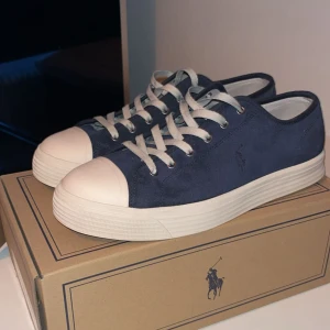   Ralph Lauren skor - Säljer ett par mörkblå sneakers från Polo Ralph Lauren med vita snören och vit tå samt sula. Skorna har en klassisk rund tå och platt sula, perfekt för en stilren look. Märkets logga syns diskret på sidan. Bara använt få tall gånger box och blåa snören medföljer!