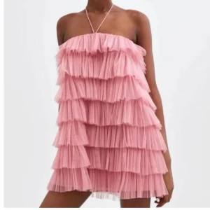 Rosa volangklänning med halterneck - Supersöt rosa kortklänning med flera lager volanger och halterneckband. Klänningen har en luftig och lekfull design som verkligen sticker ut. Perfekt för dig som vill ha något unikt och trendigt!