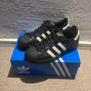 Adidas Superstar svarta - Säljer ett par Adidas Superstar, köpta på footlocker, lådan finns, vid hälen av båda skorna är fodret lite trasigt men det känns inte vid användning, kontakta gärna vid frågor!