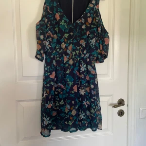 Blommig klänning från H&M Divided - Mörkblå kortklänning med blommigt mönster från H&M Divided. Klänningen har cold shoulder-detalj med korta ärmar och v-ringning fram. Perfekt för sommarens tillfällen och har en luftig passform.