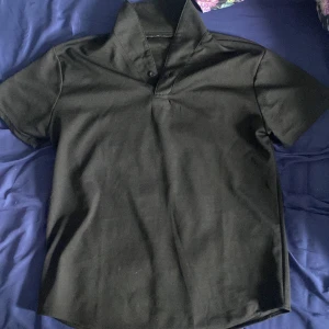 Svart piké t-shirt  - Säljer en klassisk svart piké t-shirt med krage och två knappar framtill. T-shirten har korta ärmar och är tillverkad i ett mjukt material som känns bekvämt mot huden. Perfekt för en stilren och enkel look. Storlek S men kan användas som M 