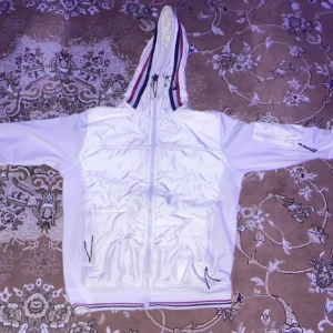 Vit jacka från Moncler nästan helt ny - Säljer en snygg vit jacka från Moncler med dragkedja och huva. Jackan har Moncler-logga på ärmen och ribbade detaljer i rött, vitt och blått. Perfekt för en stilren look.