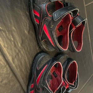 Svarta och röda sandaler från Reebok - Säljer ett par svarta sandaler från Reebok med röda detaljer och justerbara kardborreband. Skorna har en platt sula med cirkelmönster och öppen tå, perfekt för varma dagar. En strlk 23,5 och den andra 22