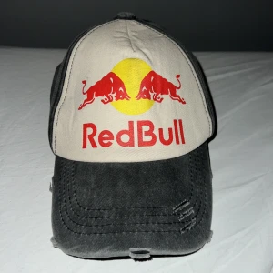 Red Bull keps med justerbart spänne - Cool keps från Red Bull med grått tyg och vit frontpanel. Framsidan har Red Bulls logga i rött och gult. Kepsen har böjd skärm och justerbart metallspänne bak för perfekt passform.