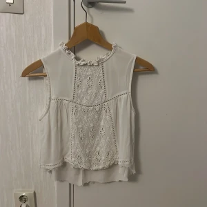 Vit ärmlös blus med spets från Indiska - Säljer en söt vit ärmlös blus från Indiska med broderade spetsdetaljer och volang vid halsen. Blusen har ett vackert mönster framtill och en lätt, luftig känsla. Perfekt för varma dagar eller lager på lager.