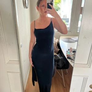 Mörkblå långklänning med slits - Säljer en elegant mörkblå långklänning med smala axelband och en hög slits på sidan. Klänningen har en tight passform och en stilren, minimalistisk design. Perfekt för festliga tillfällen när du vill sticka ut.