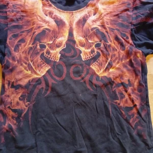 Svart t-shirt med eldiga dödskallar - Cool svart t-shirt med stort tryck av två dödskallar i eldiga orangea och röda toner över bröstet. T-shirten har korta ärmar och en klassisk rund halsringning. Perfekt för dig som gillar unika och tuffa motiv.