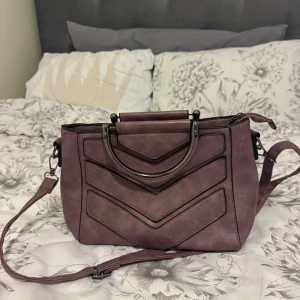 Lila axelväska med metallhandtag - Säljer en snygg lila axelväska med dekorativa metallhandtag och avtagbar axelrem. Väskan har ett modernt mönster framtill och stängs med dragkedja. Perfekt för att bära dina viktigaste saker med stil.