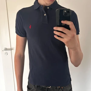 Mörkblå pikétröja från Ralph Lauren - Säljer en klassisk mörkblå pikétröja från Ralph Lauren med röd broderad logga på bröstet. Tröjan har korta ärmar, krage och knappar vid halsen. Perfekt för en stilren och avslappnad look. Slimfit storlek S