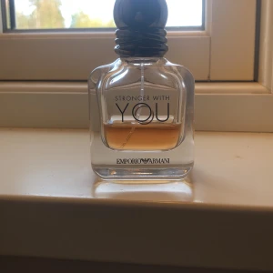 Stronger With You  - En stilren herrparfym i glasflaska med svart rund kork. Doften är Stronger With You från Emporio Armani, känd för sin varma och kryddiga karaktär. Flaskan har en transparent design med logotypen tryckt på framsidan. 15 ml av 30