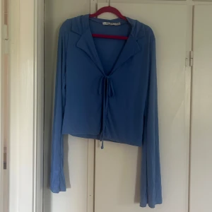 Blå långärmad topp med knytning - Säljer en blå långärmad topp från NA-KD med krage och knytning framtill. Tunt och mjukt material som ger en snygg och avslappnad look. Perfekt att styla med jeans eller kjol. Har dessvärre aldrig kommit till användning. Jättefin nu till våren och sommaren!