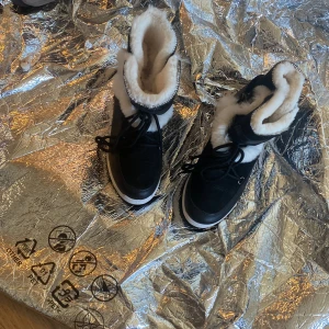 Svarta vinterboots med vit fuskpäls - Säljer ett par svarta vinterboots med vit fuskpäls runt skaftet och insidan. Skorna har snörning framtill och en tjock vit sula. Perfekta för kalla dagar när du vill hålla fötterna varma och snygga.