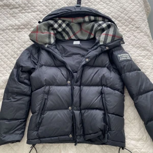 Svart pufferjacka från Burberry - Säljer den billigt just nu. Äkta svart pufferjacka från Burberry med avtagbar huva och klassiskt rutigt foder. Jackan har dragkedja och knappar framtill samt två stora fickor med dragkedja. Märkespatch på ärmen och quiltad design.