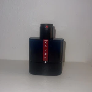 Prada Luna Rossa Carbon Eau de Toilette - Mörkblå och svart parfymflaska från Prada med röd etikett framtill. Flaskan har en modern, stilren design och är tillverkad i glas med plastdetaljer. Doften är en fräsch och maskulin Eau de Toilette.