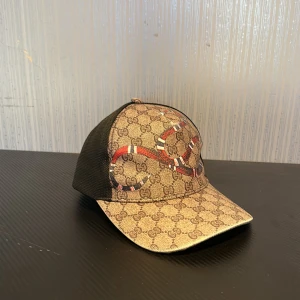 Gucci keps med orm och GG-mönster - Snygg keps från Gucci med klassiskt GG-mönster i beige och brunt, dekorerad med en broderad röd, vit och svart orm på framsidan. Baksidan är svart mesh och justerbar med grön-röd detalj.