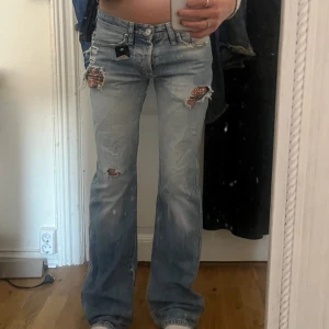 Lågmidjade bootcut/straight jeans från Dolce & Gabbana - Säljer ett par ljusblå lågmidjade jeans från Dolce & Gabbana med bootcut/straight passform. Jeansen har slitna detaljer och hål med tyg under, samt klassiska fem fickor och logga på bakfickan. 