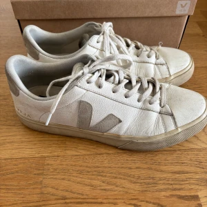 Vita sneakers från Veja - Säljer ett par klassiska vita sneakers från Veja med snörning och rund tå. Skorna har en diskret beige sula och V-logga på sidan. Perfekta för en stilren och enkel look.
