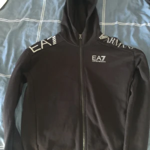 Svart hoodiejacka från EA7 Emporio Armani - Aldrig använt, fick den i present.Säljer en svart hoodiejacka från EA7 Emporio Armani med dragkedja framtill och tryckt logga på bröstet och axeln. Jackan har huva och är långärmad, perfekt för en sportig och stilren look.  