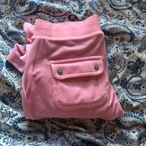 Rosa mjukisbyxor från Juicy Couture - Säljer ett par klassiska rosa mjukisbyxor från Juicy Couture i supermjuk velour. Byxorna har resår i midjan, muddar vid bensluten och en ficka med tryckknappar bak. Perfekta för en avslappnad och trendig look.