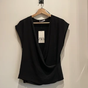 Svart omlottopp från Zara - Säljer en svart omlottopp från Zara i mjukt, lätt glansigt material. Toppen har djup v-ringning och är ärmlös med en snygg draperad effekt framtill. Perfekt att styla med jeans eller kjol för en trendig look. Helt oanvänd, köpt för 229:- säljer för 150:- 