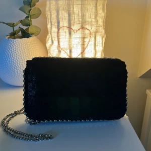 Svart axelväska med kedja från Zara - Säljer en svart axelväska från Zara med silverfärgad kedja och dragkedja. Väskan har dekorativa nitar längs kanterna och flera fack inuti för bra organisering. Perfekt storlek för det viktigaste och snygg detalj till din outfit.