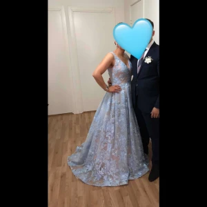 Ljusblå långklänning med spets och v-ringning - Säljer en ljusblå långklänning med v-ringad hals och breda axelband. Klänningen har spetsdetaljer. Perfekt nu inför balen eller bröllop. Köptes för 8000kr och har endast använts vid ett tillfälle och är i nytt skick och har inga defekter alls.