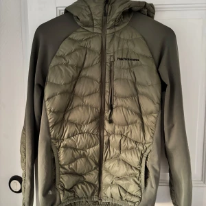 Olivgrön pufferjacka med huva från Peak Performance - Säljer en olivgrön pufferjacka från Peak Performance med huva och dragkedja framtill. Jackan har quiltade partier på bröst, rygg och huva samt släta ärmar. Perfekt för kyliga dagar och snygg till streetwear.