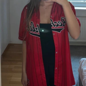 Röd basebollskjorta  - Säljer en röd basebollskjorta med ränder och broderad logga på bröstet. Skjortan har korta ärmar, knappar framtill och en klassisk basebollkrage. Perfekt för en sportig och avslappnad stil. Älskar den! Men kommer tyvärr inte till användning för mig 💗💗