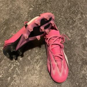 Rosa fotbollsskor från Puma - Säljer ett par snygga rosa fotbollsskor från Puma med svart och vit detalj på sidan. Skorna har snörning och mönstrad ovandel med tunna svarta och vita linjer. Perfekta för dig som vill sticka ut på planen!