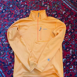 Orange fleece-tröja från Patagonia - Säljer en orange långärmad fleece-tröja från Patagonia med halv dragkedja och hög krage. Tröjan har en bröstficka med dragkedja och diskret logga på bröstet. Perfekt för lager-på-lager och utomhusaktiviteter. Varm men tunn! Inga defekter.