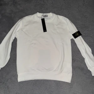 Vit sweatshirt från Stone Island - Säljer en stilren vit sweatshirt från Stone Island med klassisk rund halsringning och ribbade muddar. Tröjan har den ikoniska Stone Island-loggan på vänster ärm. Perfekt för en clean och enkel look.