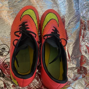 Röda fotbollsskor med svart och gul detalj - Säljer ett par röda Nike fotbollsskor med svarta skosnören och gula detaljer på ovansidan. Skorna har rund tå och dobbar för spel på gräs. Insidan är svart och grön. Perfekta för dig som vill sticka ut på planen!