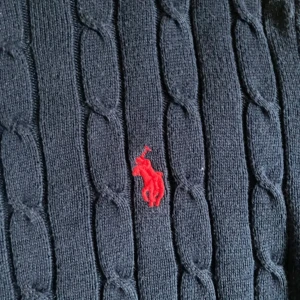  Polo Ralph Lauren - Mörkblå kabelstickad tröja från Polo Ralph Lauren med klassisk passform och rund hals. Tröjan har lång ärm och röd broderad logga på bröstet. Perfekt för en stilren och tidlös look.