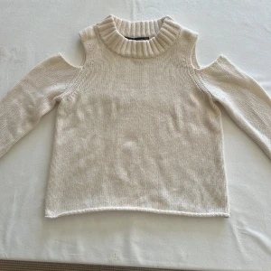 Beige stickad tröja med cut out från Karen Millen - Säljer en beige/cremevit stickad tröja från Karen Millen med coola cut out-detaljer vid axlarna och hög ribbad krage. Tröjan har lång ärm och är perfekt för kalla sommarkvällar. 