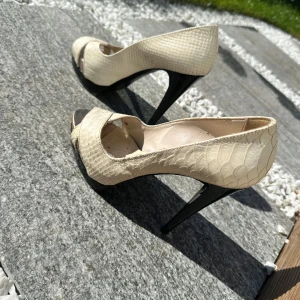 Beige pumps med ormskinnsmönster och öppen tå - Säljer ett par eleganta beige pumps från SORETTI med hög klack och öppen tå. Skorna har ett stilrent ormskinnsmönster och är tillverkade i syntetmaterial. Perfekta för dig som vill sticka ut med en klassisk men trendig look.