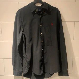 Svart skjorta från Ralph Lauren - Säljer en svart långärmad skjorta från Ralph Lauren i slim fit-modell. Skjortan har klassisk krage, knappar framtill och en liten röd broderad logga på bröstet. Perfekt för dig som gillar stilrena plagg med snygga detaljer. Skulle säga att den passar runt 185cm. Priser går att diskutera vid snabb affär