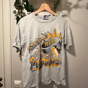 Grå Pittsburgh Penguins NHL t-shirt från Trench - Säljer en grå vintage t-shirt från Trench med stort Pittsburgh Penguins NHL-tryck i svart, gult och vitt på framsidan. T-shirten är i 100% bomull och har normal passform med korta ärmar. Perfekt för hockeyfans! På bild tre ser man att det finns lite vit färg som jag inte fått bort, ingenting man tänker på när den är på