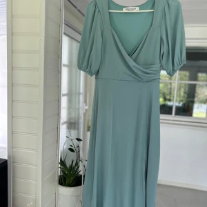 Ljusblå långklänning med omlott och puffärm - Säljer en ljusblå långklänning med omlott framtill och v-ringning. Klänningen har korta puffärmar och ett mjukt, följsamt tyg som ger ett elegant fall. Perfekt för somriga tillfällen eller studentfirande.